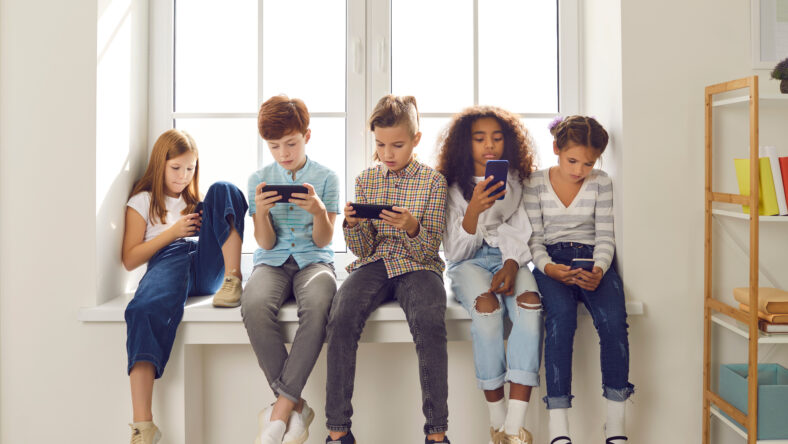 Group of children of different nationalities sit in a row on the windowsill and with lack of emotion play online games or read social networks on mobile phones. Technologies that spoil childhood.