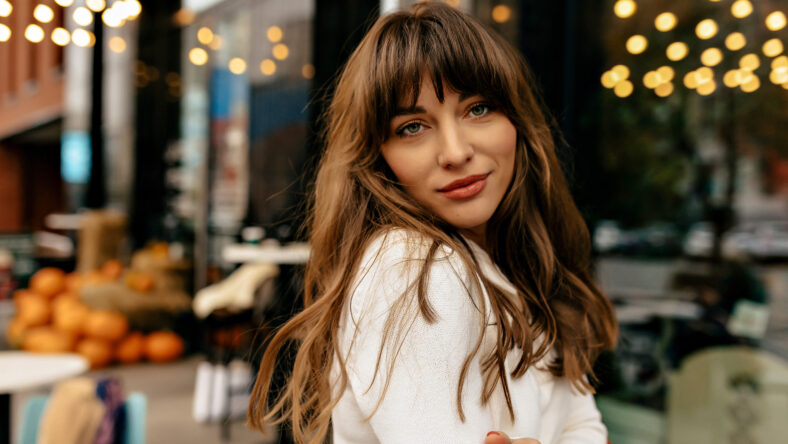 Stylish pretty girl with dark wavy hair wearing knitted white sweater posing at camera on city lights. She is smiling and crossed arms on waist