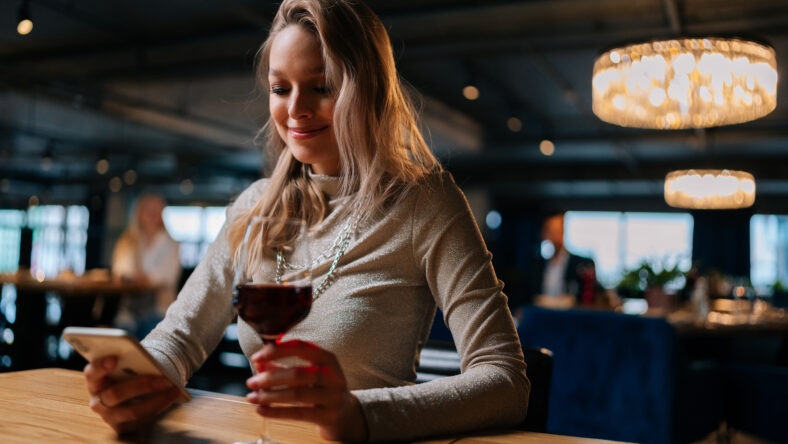 Low-angle view of smiling blonde young woman using smartphone, typing online message sitting at table holding in hand glass of red wine at restaurant. Happy pretty lady chatting with boyfriend.
