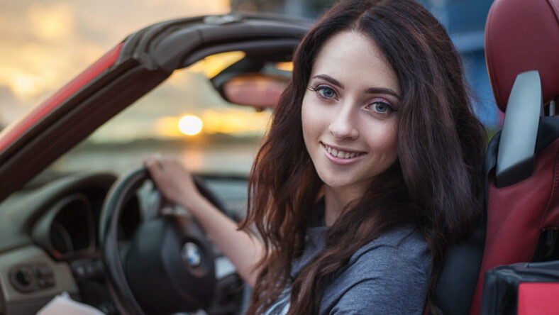 Pretty young brunette woman driving luxury red cabriolet car at the sunset