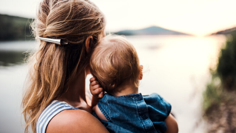A rear view of mother with a toddler daughter standing outdoors by the river in summer.