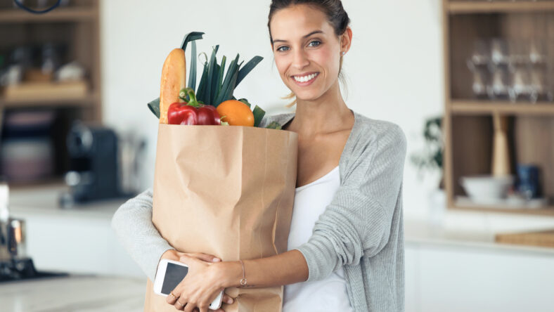 Portrait of beautiful young woman looking at camera while holding shopping bag with fresh vegetables in the kitchen at home.