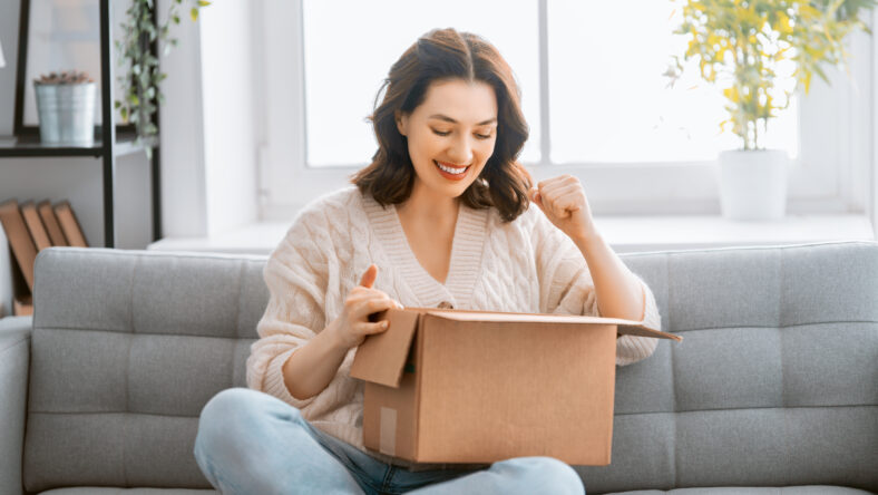 Beautiful young woman is holding cardboard box sitting on sofa at home.