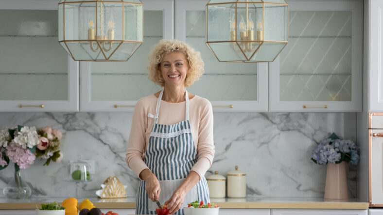 Happy pretty middle aged cooking blogger woman in apron cutting fresh vegetables in modern kitchen interior, standing at table, looking at camera, smiling. Healthy food eating. Indoor portrait