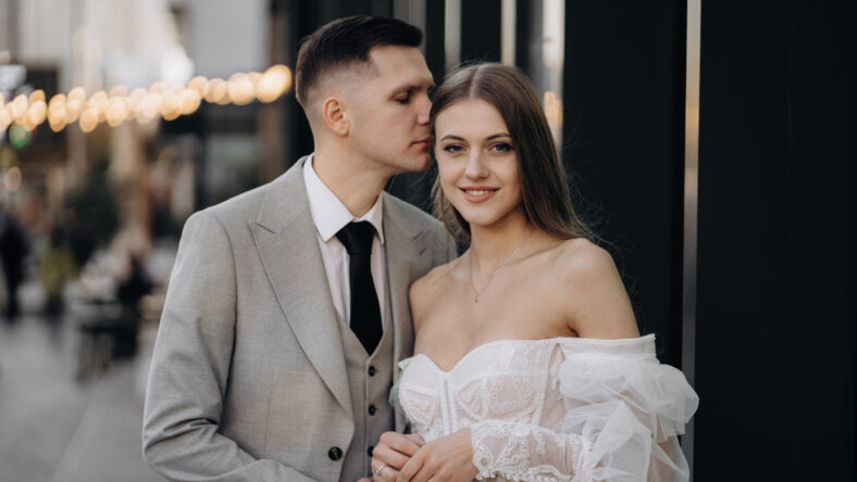 Front view of happy couple having wedding phtoshoot outdoors. Handsome groom kissing beautiful bride, woman looking at camera, smiling. Concept of marriage and happiness.