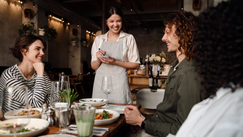 Young smiling waitress wearing apron serving group of cheerful young friends in restaurant
