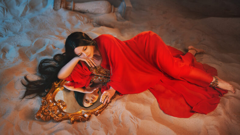 woman lying on sand and looking into a mirror while wearing a red dress