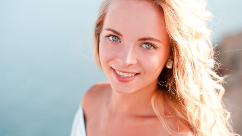 Smiling blonde girl posing over sea background outdoors. Looking at camera. 20s.