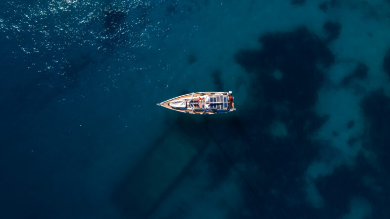 Directly above, a single sailboat rests on deep blue water. Sun glints on the sea, reefs tint patches, and the hull casts a long diagonal shadow.