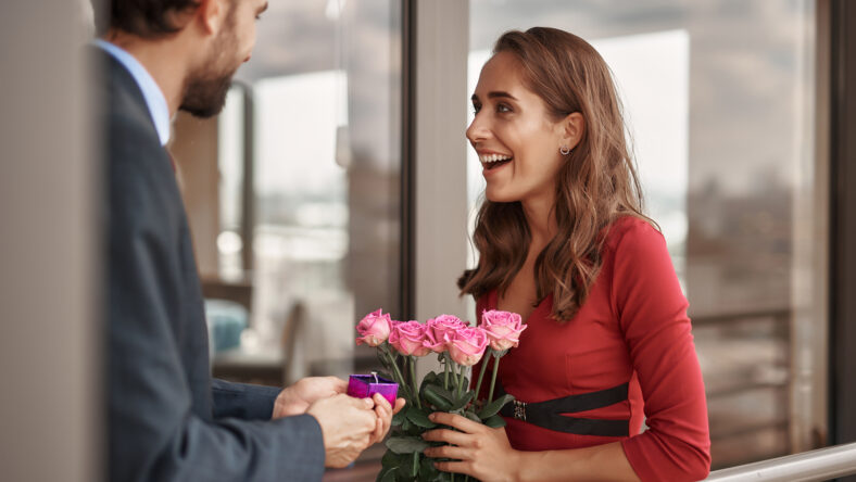 Concept of surprising engagement. Waist up portrait of young male holding box with ring and making marriage proposal to his happy lover on balcony
