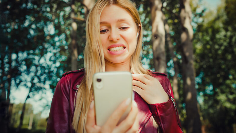 shock shout beautiful young pretty girl looking at the screen display of mobile phone reading watching fail bug news chat sale outdoor in the park reading and having communication with teeth