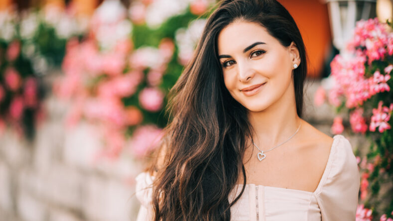 Close up portrait of beautiful young woman with long shiny hair, posing outdoor in pink flowers