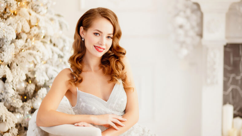Young beautiful woman smiling and happy looking camera, hair styling makeup, white silver color on background of Christmas tree and fireplace bokeh.