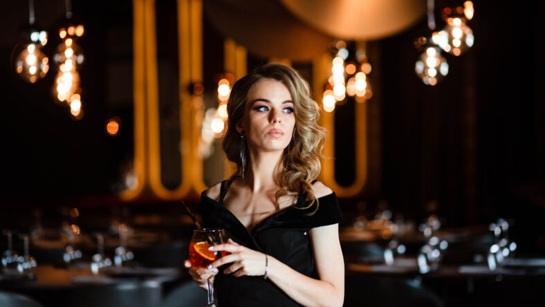Beautiful blonde, young girl holding an aperol spritz. Cocktail aperol spritz in a glass. Portrait of a young girl in a restaurant. Portrait of a girl. Young girl in a restaurant with a glass.
