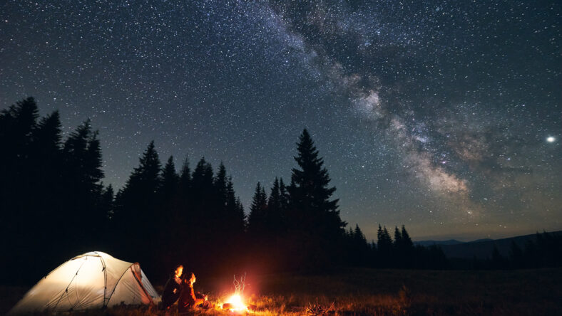 Side view of loving couple sitting near bright burning campfire and tent, enjoying beautiful camping night together under dark sky full of shiny stars and bright Milky Way, warm summer night.