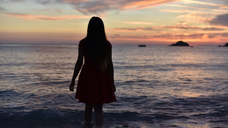 woman in dress walking away to sea on sunset