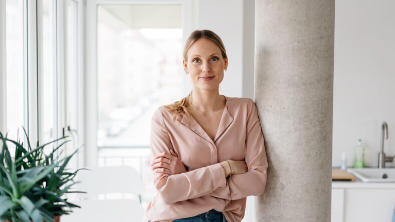 Friendly smiling young woman relaxing in an apartment kitchen leaning against an interior pillar with crossed arms looking at camera in a high key portrait with copyspace