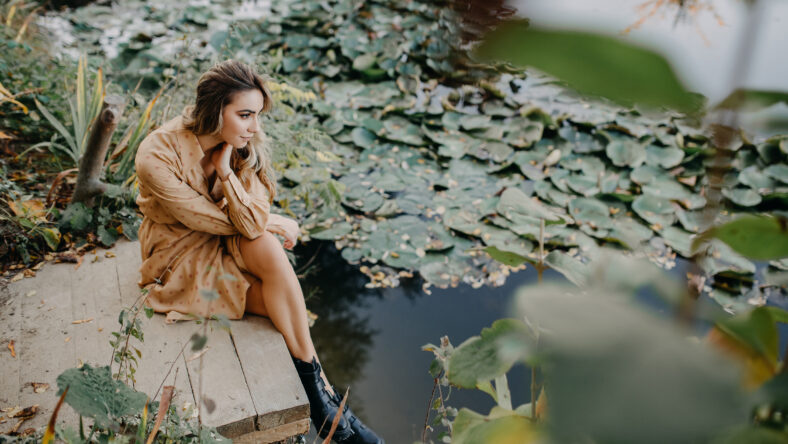 Young woman wearing a maxi chiffon dress sitting on a wooden pier by a pond with water lilies.