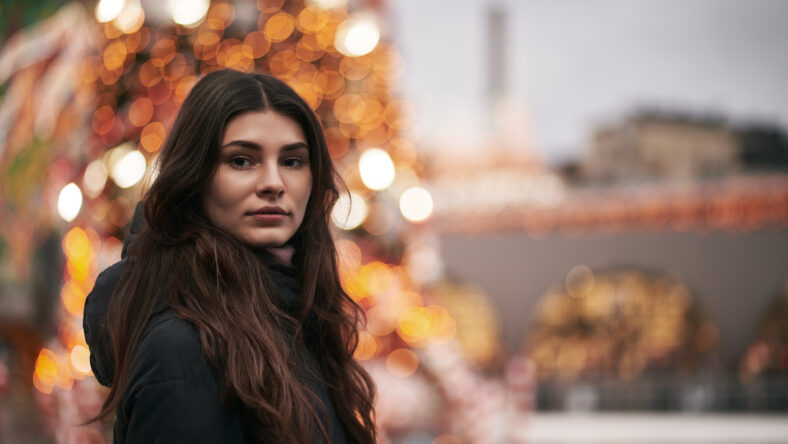 A beautiful and young girl looks into the camera. Christmas city decoration blurred in the background. Cute brunette faced to the camera in the downtown before New Year party