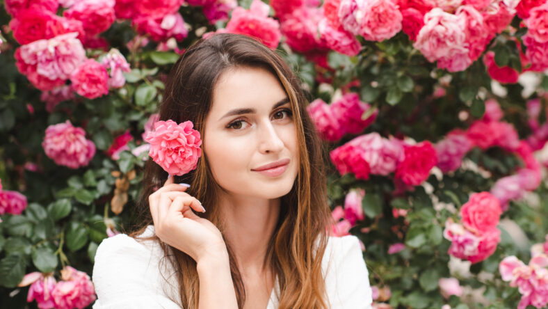 Smiling beautiful woman with long blonde hair and rose flower over blooming nature background outdoor. Attractive female with healthy skin and floral hairstyle. Springtime.