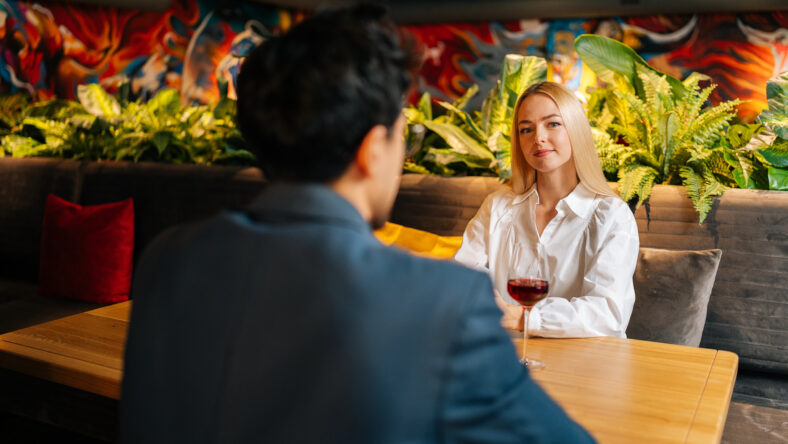 View from back of unrecognizable young man to beautiful blonde woman sitting at table with glass of red wine having festive dinner at restaurant. Happy loving couple enjoying talking together.