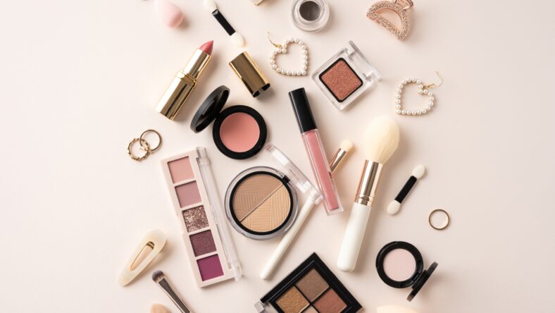 Flat lay of assorted makeup products including lipstick, blush, and brushes on a beige background