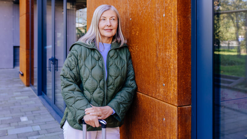 Elderly woman with long hair standing outside modern building holding suitcase relaxed in casual outfit with green jacket, suggesting travel or vacation. Adventure, leisure, and freedom in older age.