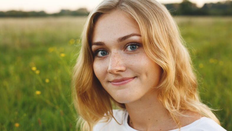 A portrait of beautiful young blue-eyed girl with light hair having charming smile and dimple on her face looking into camera standing over green background having nice time relaxing outdoor in summer