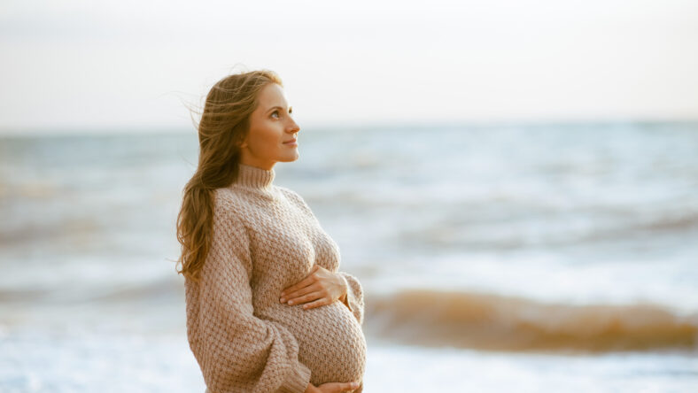 Smiling pregnant woman 25-29 year old holding tummy wearing knitted dress posing over sea outdoors. Autumn season. Prenatal lifestyle.