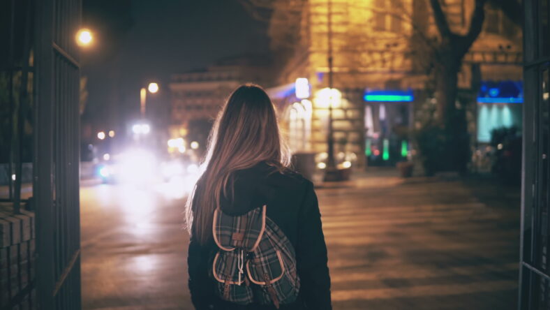 Back view of young brunette woman walking late at night in Rome, Italy city centre. Girl with backpack turns and see the Colosseum., the main sight of the city. Female spending evening alone.