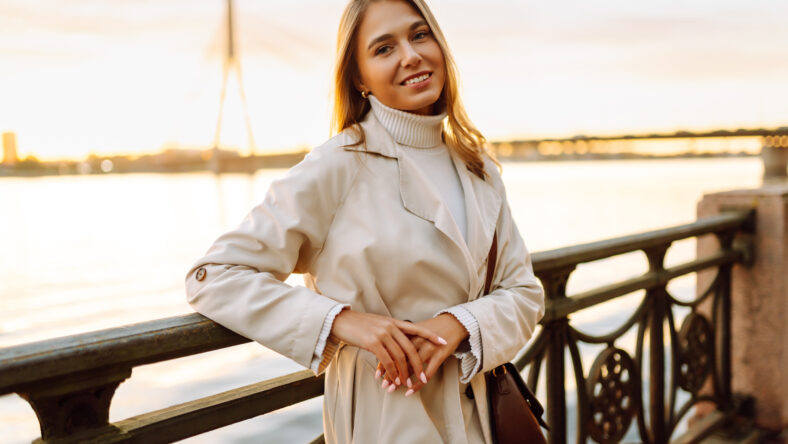 Portrait of young woman in elegant coat posing on embankment in sun rays. Beautiful woman enjoying sunset near river. Beauty, style and walks concept.