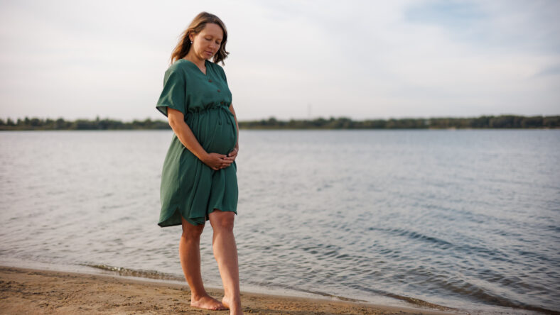 Pregnant girl in green dress walks with dog on lake shore. Owner walks Jack Russell terrier on sea shore. Pet training. Portrait of pregnant woman.