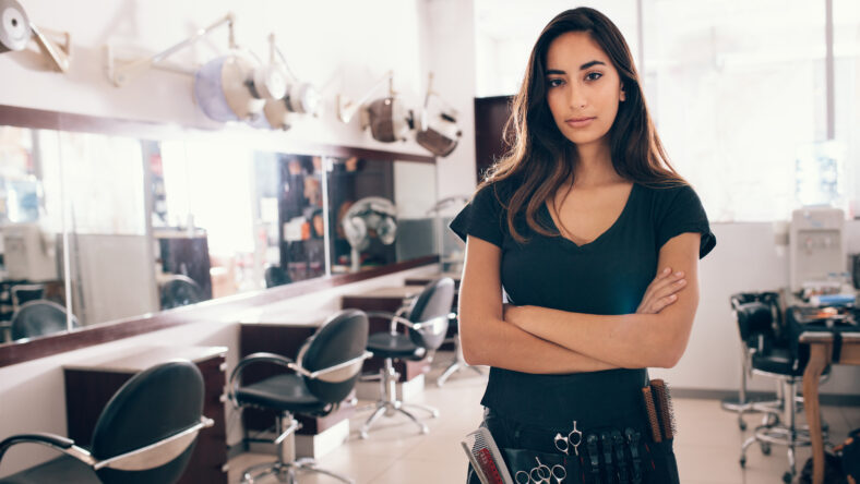 Woman standing with hands folded in her salon. Professional hairdresser with all the hairdressing accessories tied to her waist.