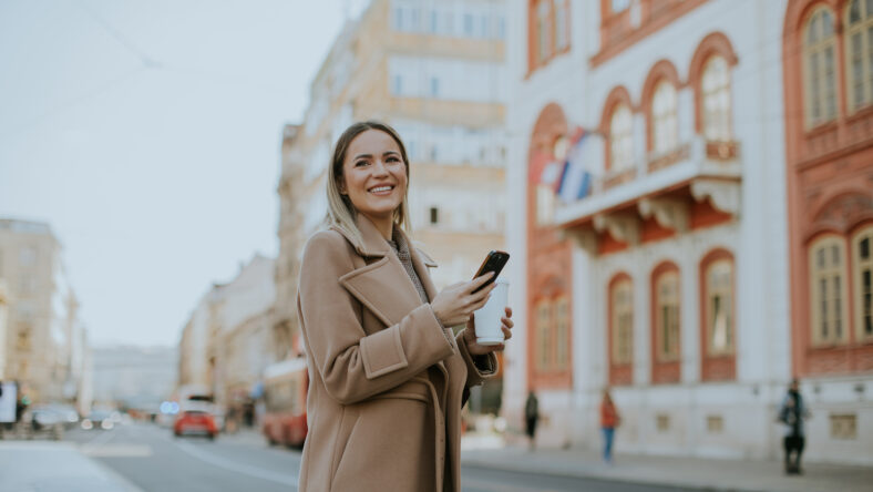 A cheerful woman stands on a city street, holding her smartphone and enjoying the sunny day. Colorful buildings line the street, creating a lively and inviting atmosphere.