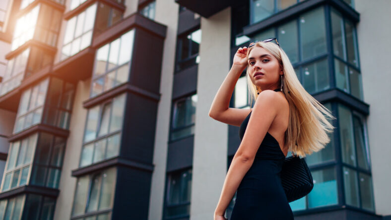 Portrait of young blonde woman wearing stylish clothing and accessories by modern business center in city. Model turning around