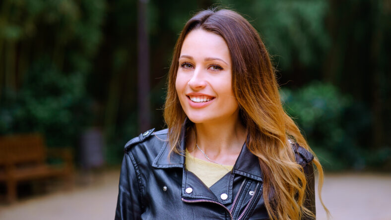 Attractive young woman charmingly smiling and looking at camera while standing on blurred background of park