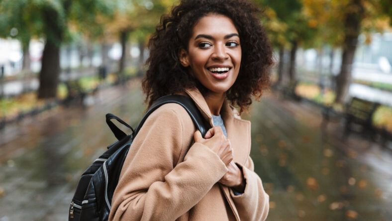Beautiful young african woman wearing coat walking outdoors at the park, carrying backpack