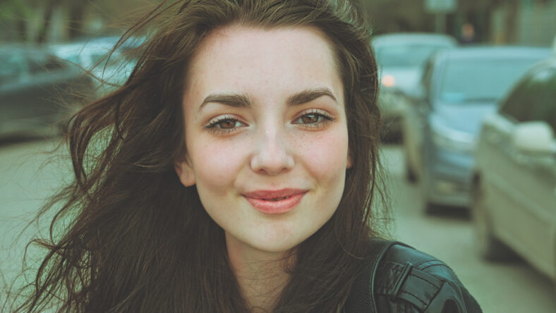Happy girl posing outside in the street with parked cars on background looking at camera with windy hair