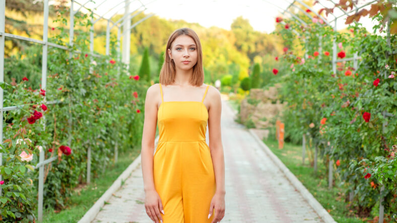 Cute Woman smiling standing on a way path posing in the garden near green bushes with pink roses. Girl wearing a yellow jumpsuit in half body length looking at you, camera.