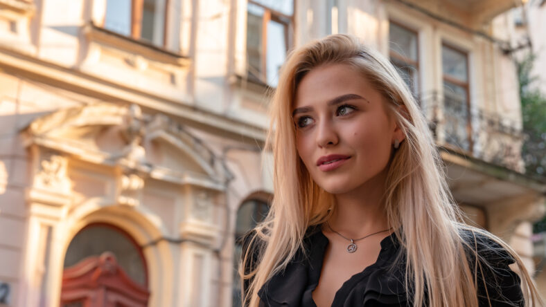 Waist up portrait of attractive stylish blonde woman with loose hair walks among old city center, summer sunny day