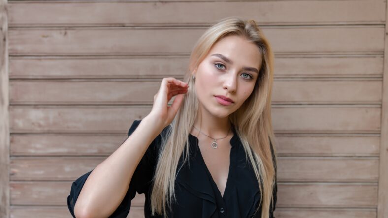 Waist up portrait of the blonde girl with blue eyes, dressed in black dress, holds hand near her face and looking at the camera, wooden background, outdoor photo