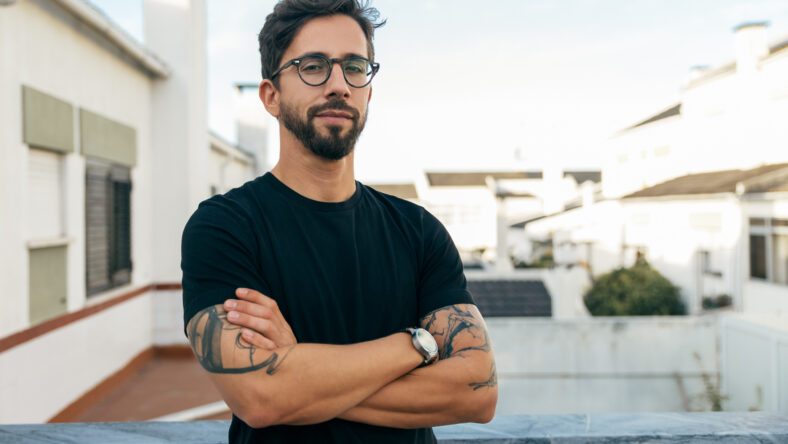 Confident stylish guy with tattoos posing on apartment balcony or terrace. Young man in glasses standing outside with arms crossed and looking at camera. Male portrait concept