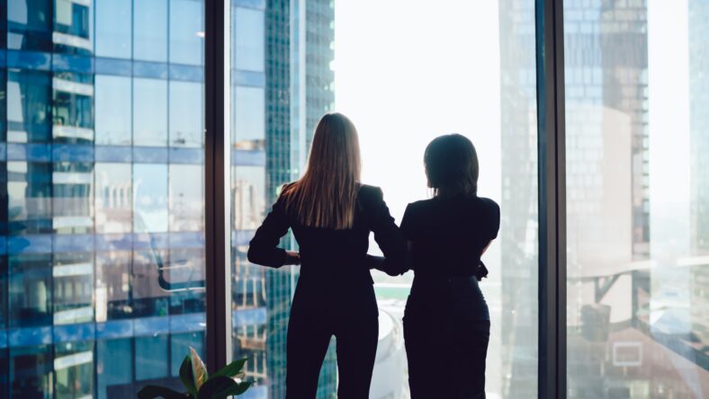 Back view of female colleagues in formal wear standing near window looking at modern exterior of skyscrapers in business center, prosperous women standing together planning prosperous future success