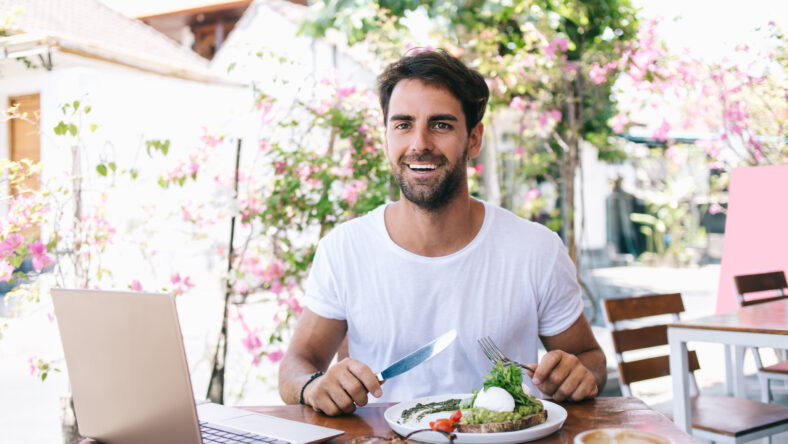 Half length portrait of cheerful caucasian male freelancer making break for lunch enjoying healthy food at cafe terrace, smiling man looking at camera eating salad on brunch holding fork and knife