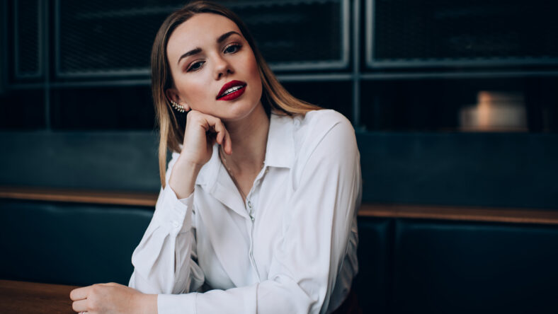 Thoughtful stylish young female in white shirt with fancy makeup sitting at table in contemporary dark place and looking at camera