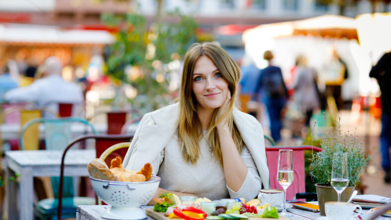 Young beautiful woman drinking coffee and having breakfast while waiting for friend. Happy alone woman in outdoor cafe or restaurant on sunny summer or early autumn day