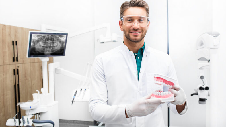 Confident male dentist standing in his modern clinic. Dental hygienist wearing protective glasses smiling at the camera