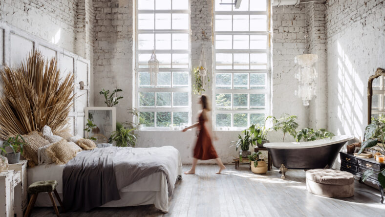 Silhouette of young woman walking at home with interior design in bohemian style against loft brick wall with large windows. Dreamy female standing between cozy bed in bedroom and bathroom bath