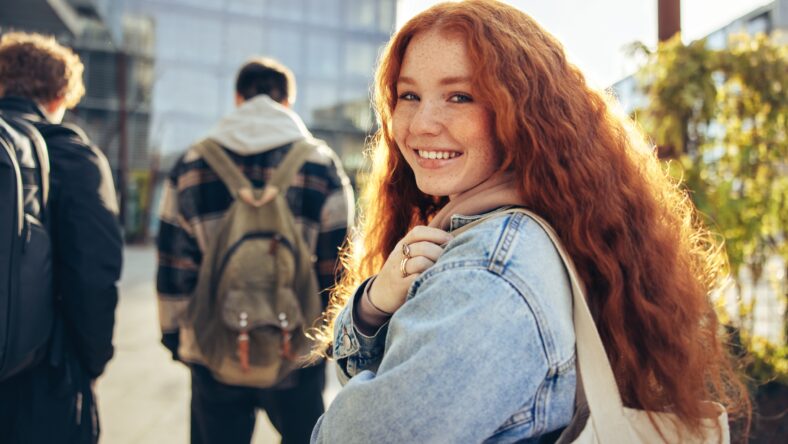Girl looking back and smiling in college campus. Female student with red dyed hair going for her lecture.