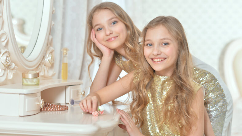 Portrait of cute smiling twin sisters in glittering golden dresses sitting near dressing table